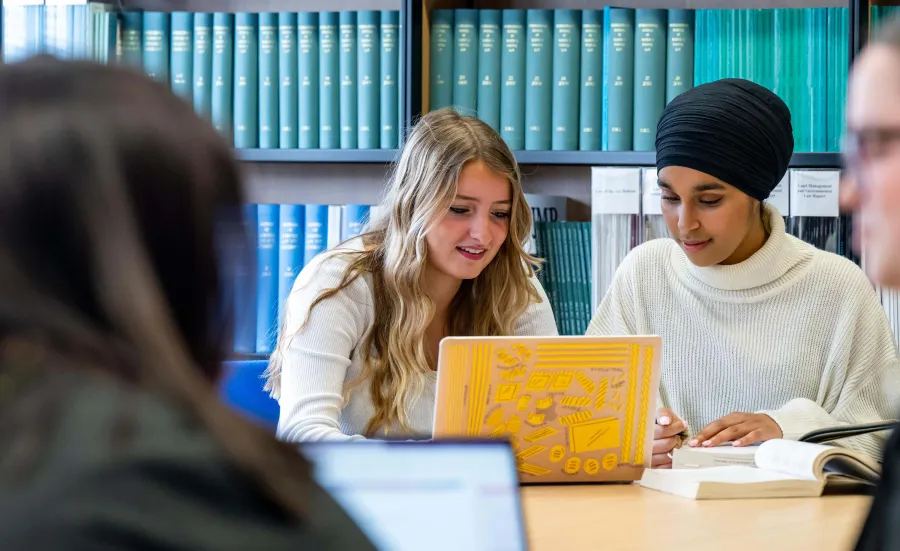 Two students discussing work in a library with a yellow laptop.