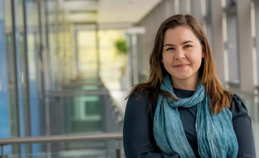 A young woman in a dark blouse and blue scarf smiles confidently in a modern university hallway.