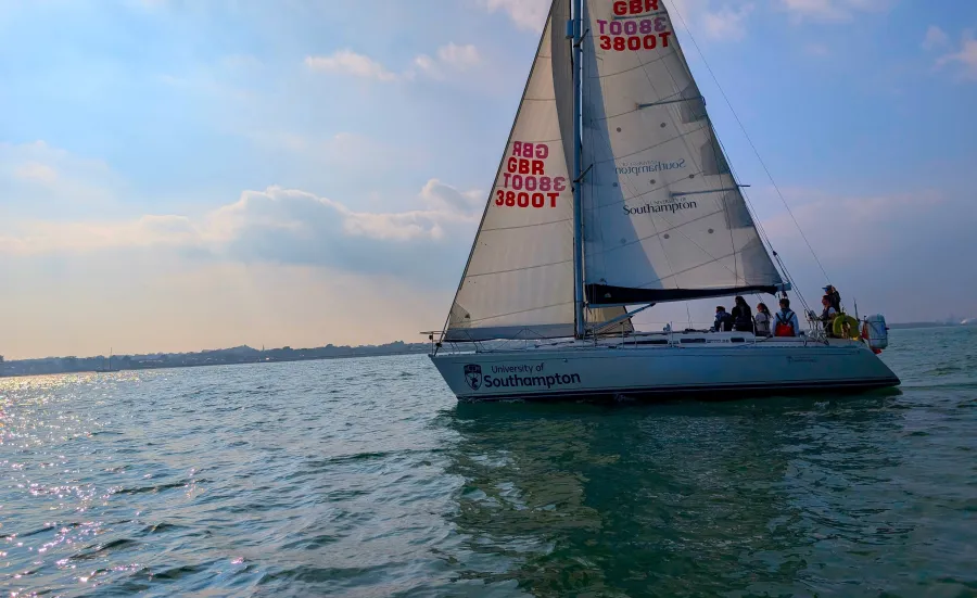 A yacht with 2 sails and a training crew sailing out on the water. The  sky has clouds and an orange eveningglowsky