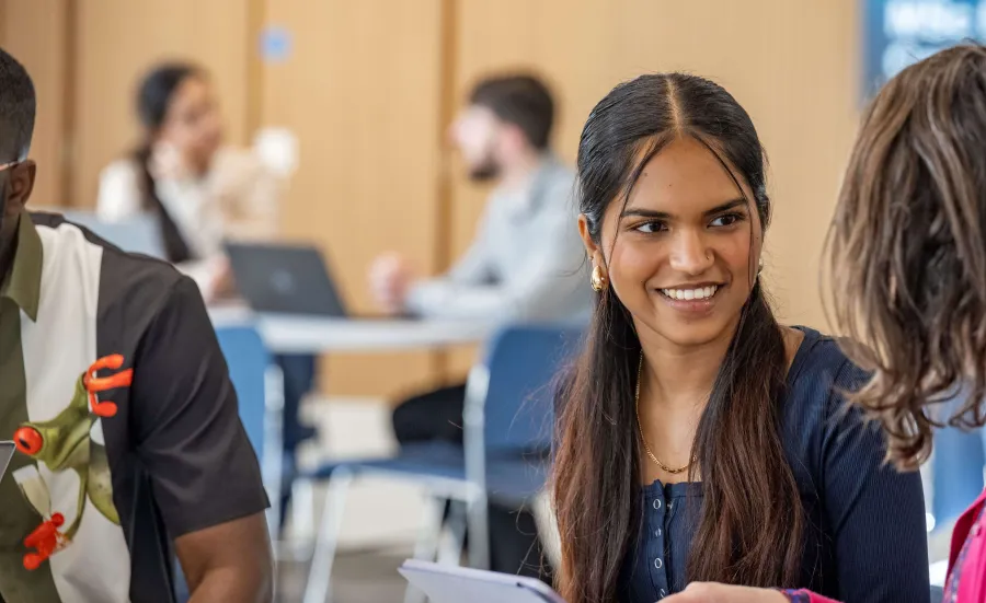 An environmental science undergraduate student talking with a tutor and referring to information on a tablet. They are sitting around a desk with other students in the background.  