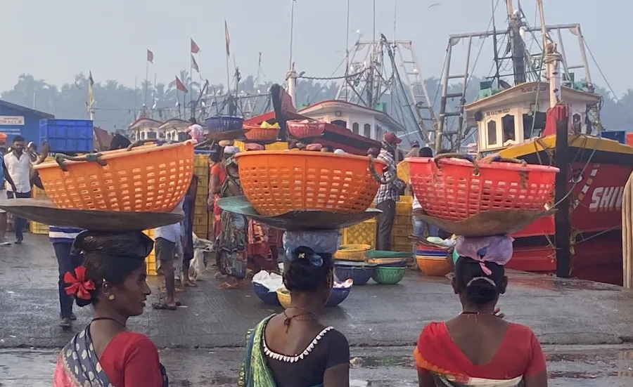 Three women at a ship harbour carrying baskets of fish on their heads.