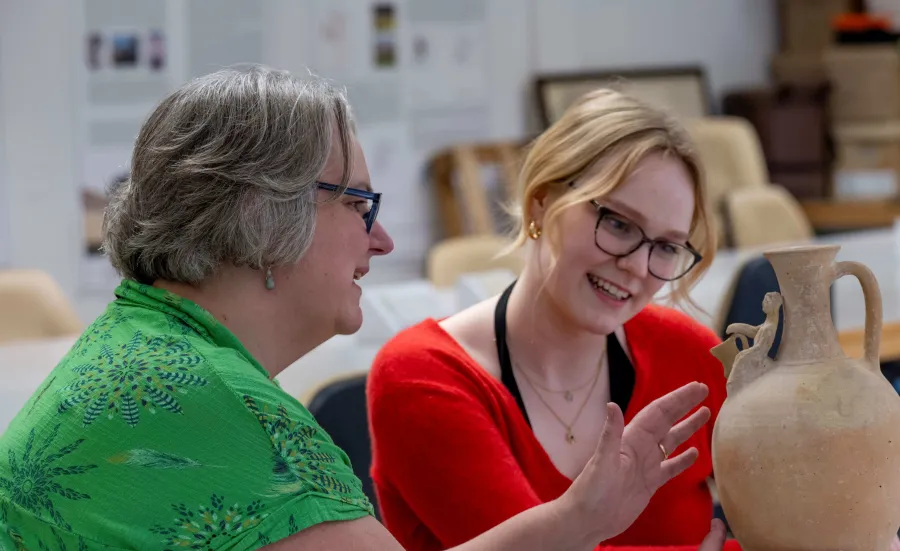  Archaeology students examining an ancient pottery jug in the Archaeology lab. There is a small human form with their hand leaning on the spout.