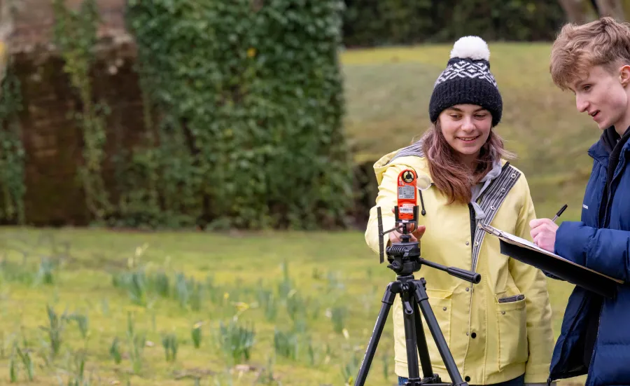 Two geography students outside using a theodolite to measure the ground. One student is recording information on a clip board.
