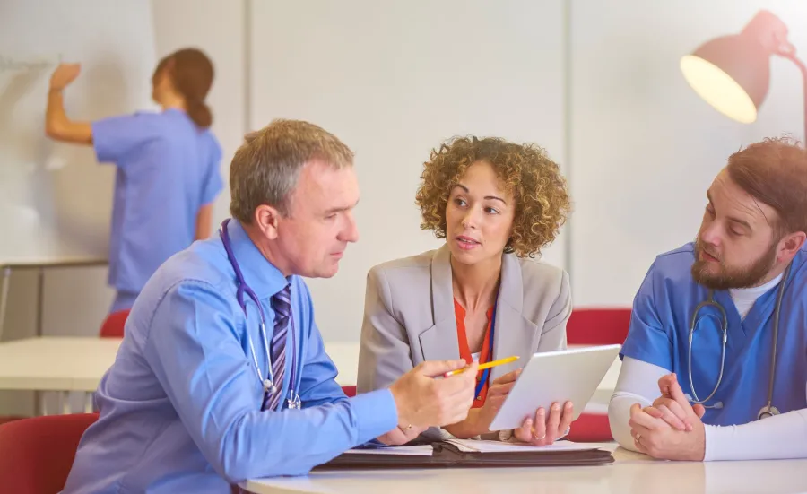 People in a meeting looking at a tablet. One of the people is wearing hospital scrubs. There is someone in the background writing on a whiteboard.