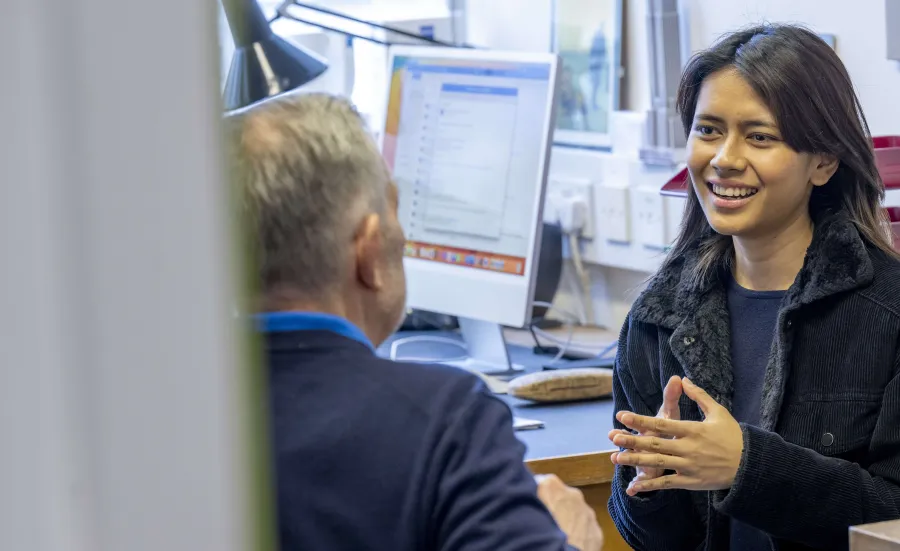 A Philosophy and Politics student is talking to one of the lecturers in an office. The office desk has a computer screen and angle poise lamp. 