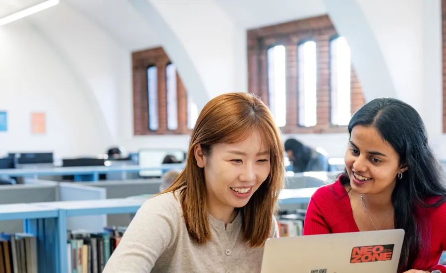Two students sitting at a table in a library. They are smiling and looking at a laptop.