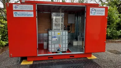 A red container unit with open doors showing an IBC tank and electrical components inside. Logos for "The Faraday Institution," "University of Southampton," and "soLead energy" are on the exterior
