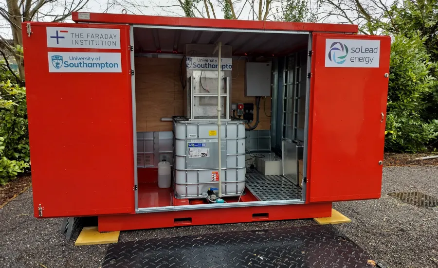 A red container unit with open doors showing an IBC tank and electrical components inside. Logos for "The Faraday Institution," "University of Southampton," and "soLead energy" are on the exterior