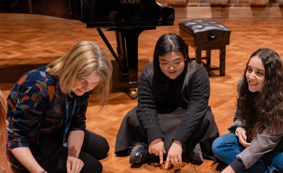 Music Education students sitting in a circle on the wooden floor of the Turner Sims hall. They are pointing to and discussing music sheets in front of them.