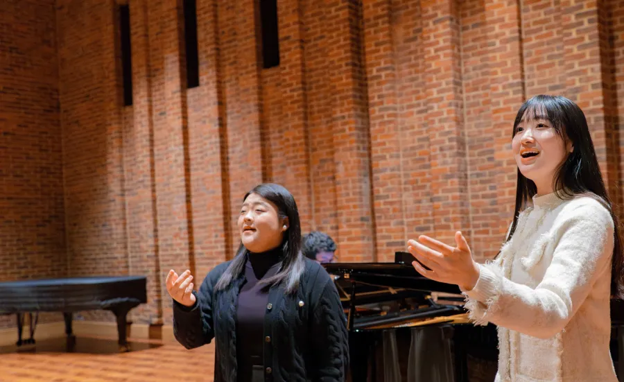 Two music students singing in the Turner Sims hall, while another student accompanies them on a grand piano.
