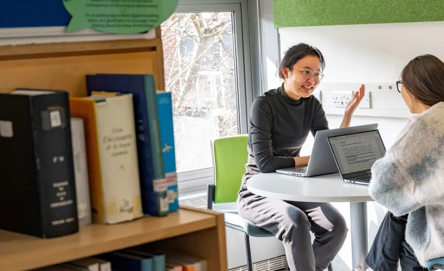 Two Translation and Professional Communication Skills masters students sitting at a table chatting. They both have a laptop on the table in front of them.