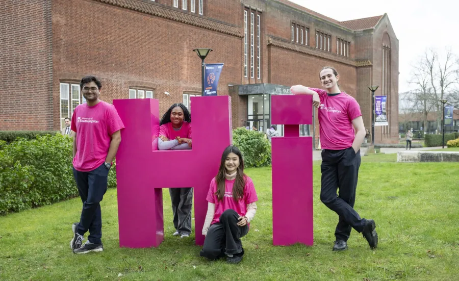 Four students leaning on large pink letters spelling Hi on the grass outside the Hartley library.