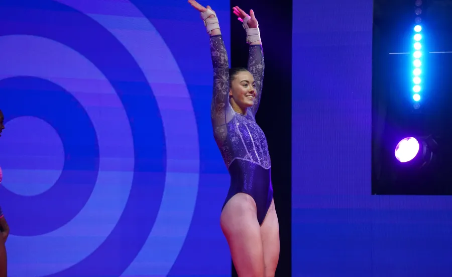 Elite athlete Emily Burke in a purple and silver leotard stands with her arms raised, smiling beneath stage lights after completing her routine.