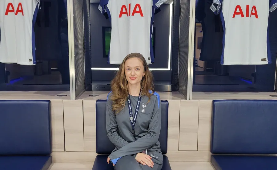 Emma Green wearing a Tottenham Hotspur tracksuit, is sitting on a chair with 3 Tottenham Hotspur shirts hanging behind her.