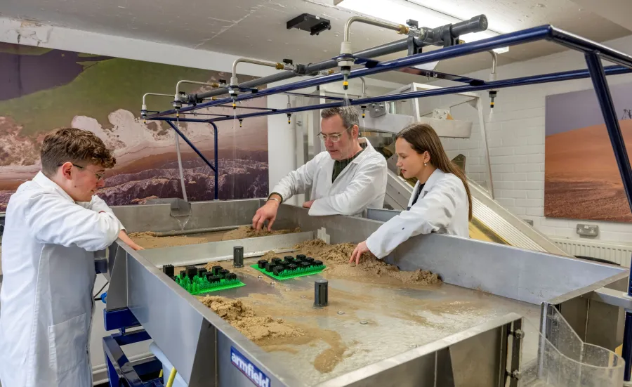 Three people in lab coats work at a sand-filled table with various objects and overhead nozzles, set against a backdrop of landscape images.