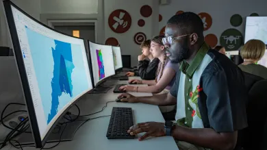 Students working at computers with large, curved monitors displaying colorful maps in a dimly lit room