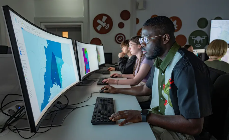 Students working at computers with large, curved monitors displaying colorful maps in a dimly lit room
