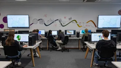 Students sit at desktop computers in a tech-enhanced room with vibrant wall graphics and digital maps on large screens