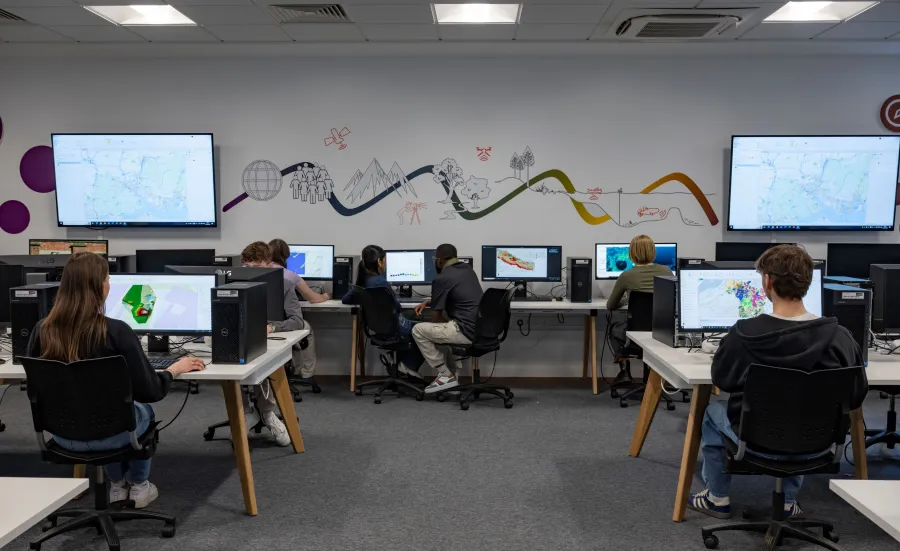 Students sit at desktop computers in a tech-enhanced room with vibrant wall graphics and digital maps on large screens