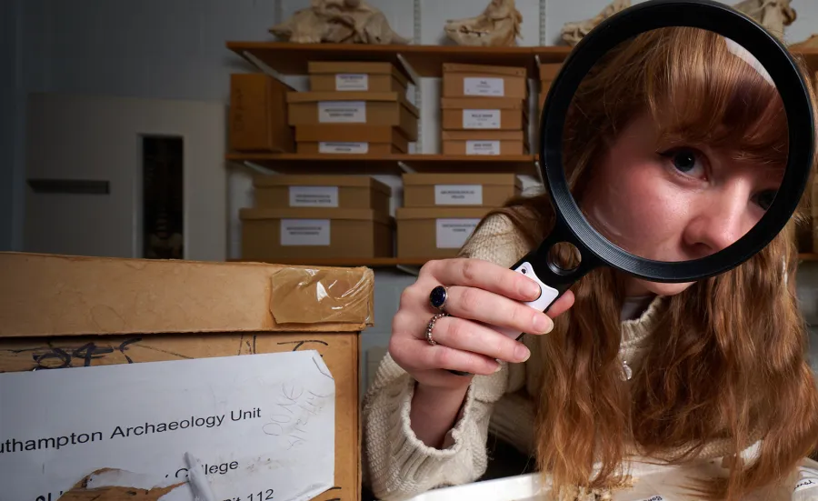 An archaeology student in an archives room, using a magnifying glass to inspect a tray of bones and fossils. The student has red hair and is wearing a cream-coloured knitted sweater. There are more fossils and bones in the background, as well as shelves filled with boxes.