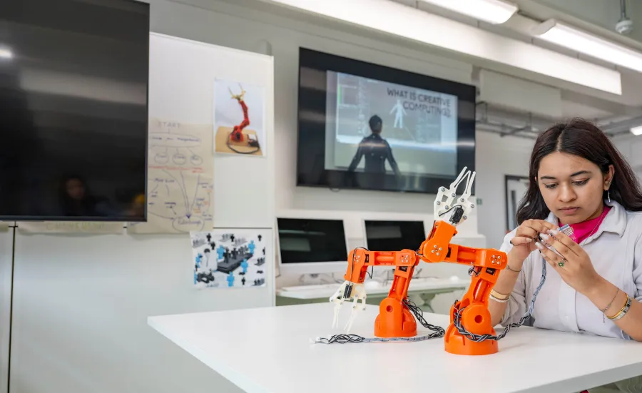 Creative computing student is assembling components beside an orange robotic arm in the creative computing lab.