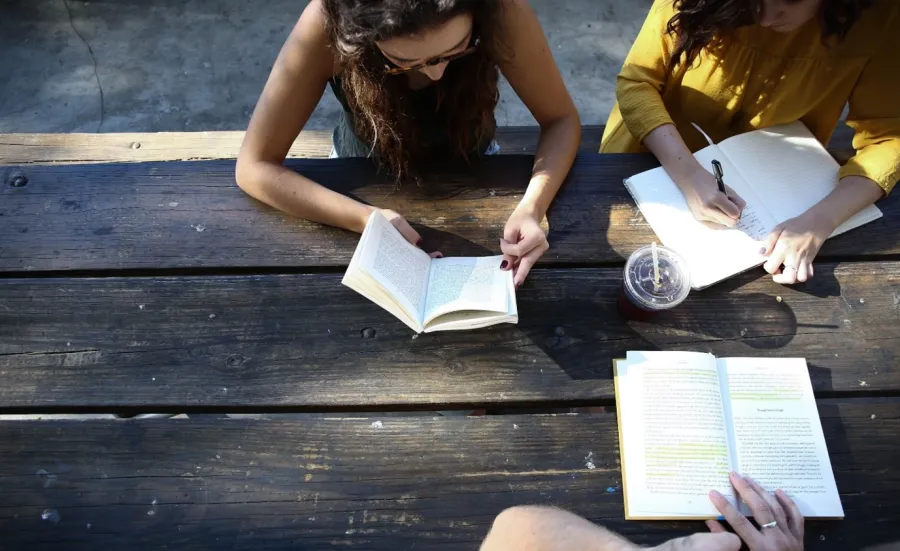 Three people gathered around at a picnic table while reading books and writing notes
