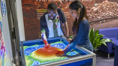 Two students exploring an interactive topographic sandbox display with projected contour and elevation colours.