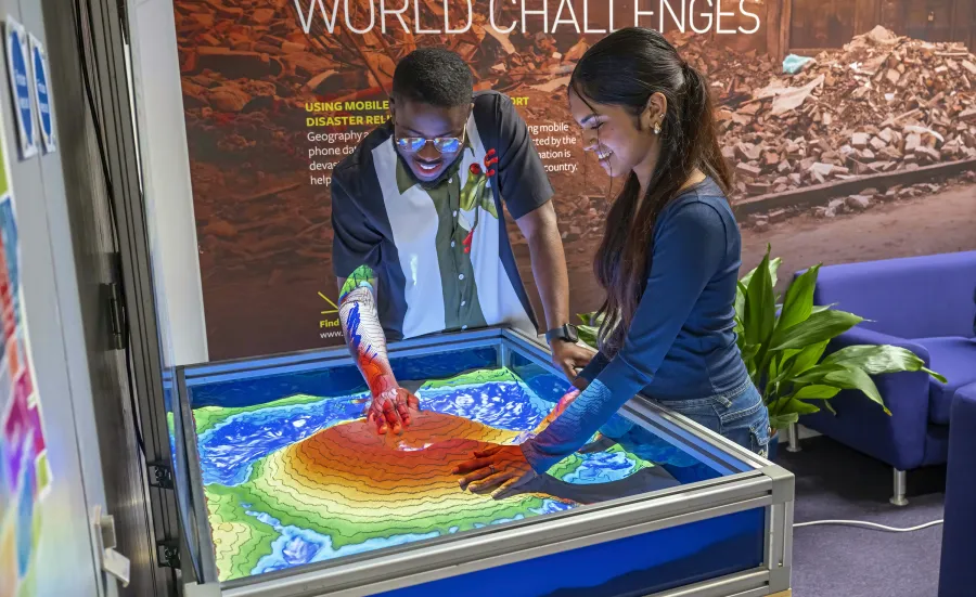 Two students exploring an interactive topographic sandbox display with projected contour and elevation colours.