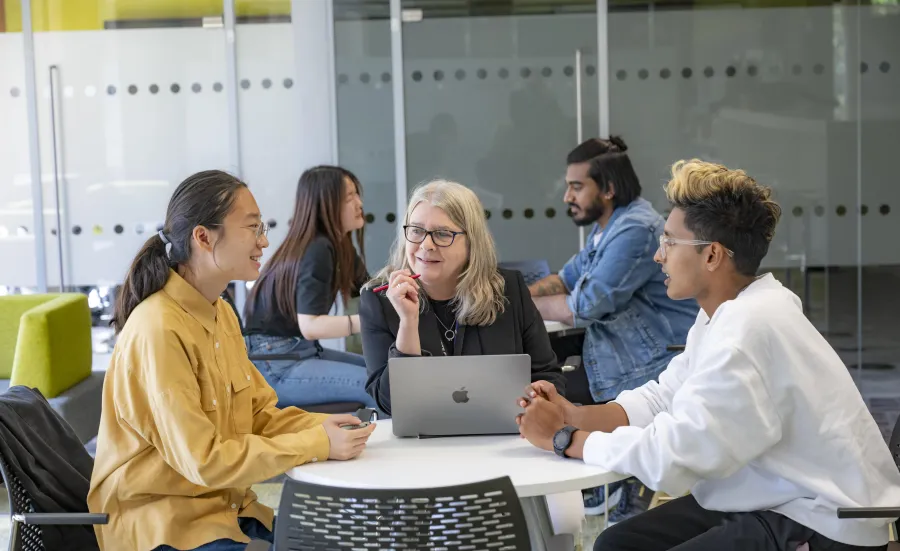 Three people sat a desk talking. The person in middle has a laptop open on the desk.