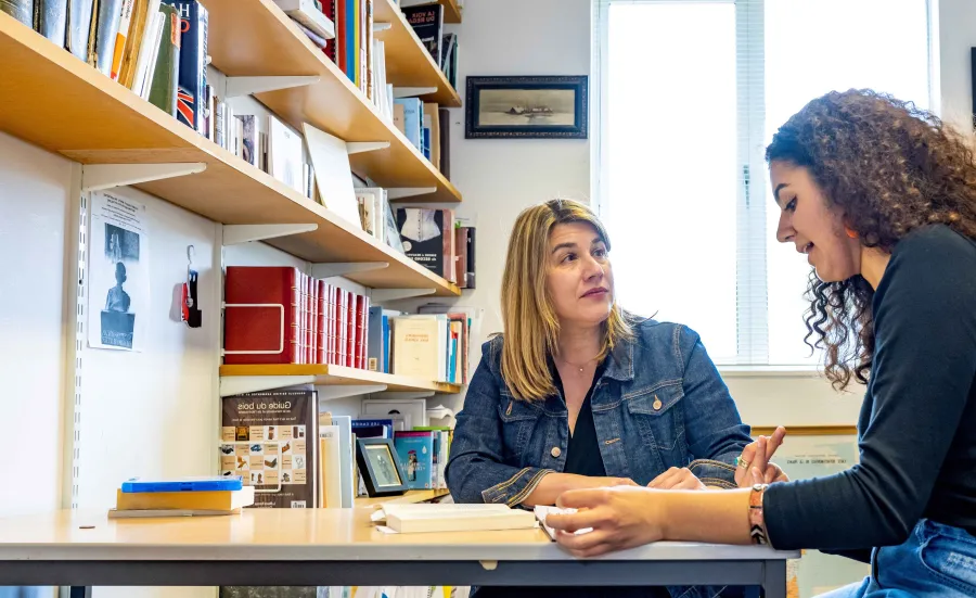 A modern language student sitting at a desk with her tutor. They are in an office with shelves of books and pictures on the walls. 