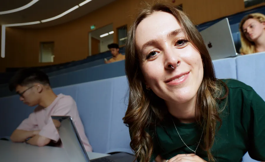 A student smiling while sat in a lecture theatre, with a laptop open on the desk in front of them.