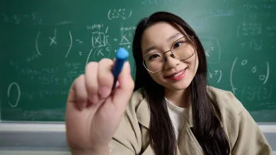 A student using a white marker pen to write a mathematical equation on a glass surface. They are wearing glass and a beige jacket, and they are stood in front of a blackboard which also features a mathematical equation.
