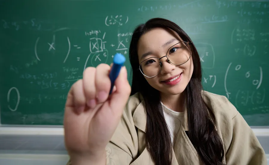 A student using a white marker pen to write a mathematical equation on a glass surface. They are wearing glass and a beige jacket, and they are stood in front of a blackboard which also features a mathematical equation.