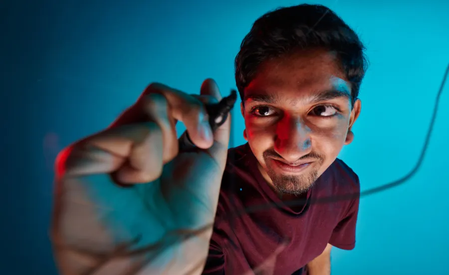 A student using a black marker pen to draw a line on a glass screen. The student is wearing a dark red t-shirt and is positioned in front of a blue background.