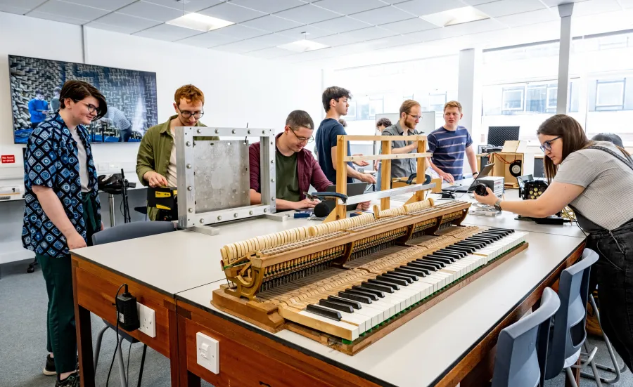 A group of students sits around a long table in a classroom, where a piano keyboard has been placed for an experiment.
