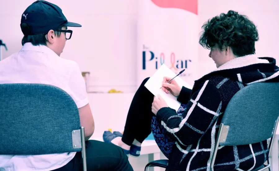 Two young adults sitting with their backs to the camera. One has their feet up on a table and is writing on a note pad resting on their knee. The other has a baseball cap on and is watching the person writing. there is a Pillar logo on the wall.