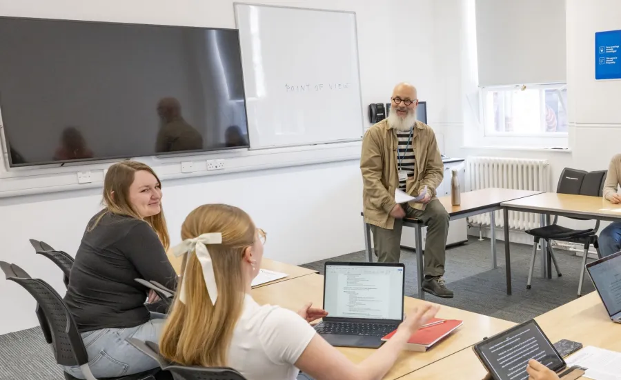 Alt text: A teacher sits on a desk holding papers while three students face them, one using a laptop. A whiteboard behind the teacher displays the phrase "POINT OF VIEW."