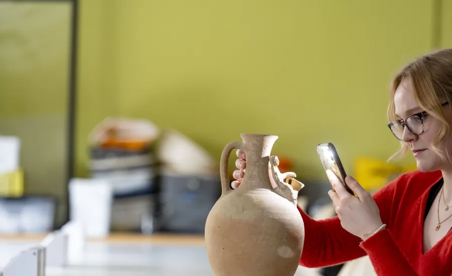 An ancient history student is examining a pottery jug using her mobile phone torch. She is looking at a small carving of a figure around the neck of the jug.
