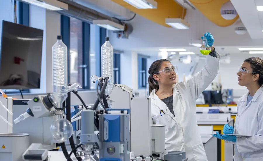 Two students in white coats in the newly refurbished chemistry labs with distillation glass ware set up. They are wearing safety glasses and looking up at a flask containing a yellow liquid. 
