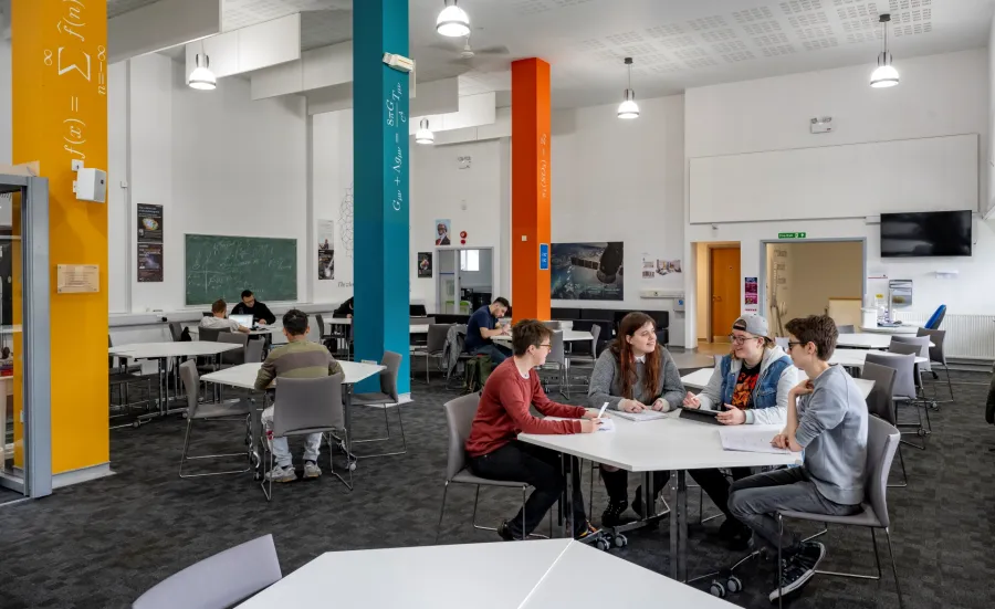 A study and social space on a university campus. In the foreground, there is a group of four students sat around a table. There are more tables with students in the background, and the pillars around the room have mathematical equations written on them.