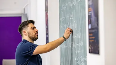 A student writing a mathematical equation on a blackboard, in a study and social space on a university campus.