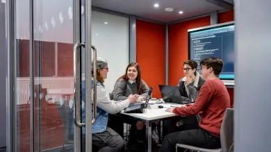 Four people sat around a table in a small meeting room. Two of the people have laptops open on the table, and there is a TV screen mounted on the wall showing code. The walls of the meeting room are red, and the glass door to the room is open.