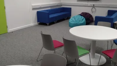 An empty room with sofas, beanbags and round tables with chairs surrounding them. There is also a TV mounted on the wall.