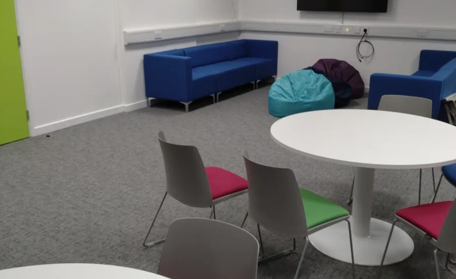 An empty room with sofas, beanbags and round tables with chairs surrounding them. There is also a TV mounted on the wall.