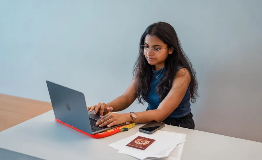 A student sat a desk with a laptop, mobile phone, passport and documents.