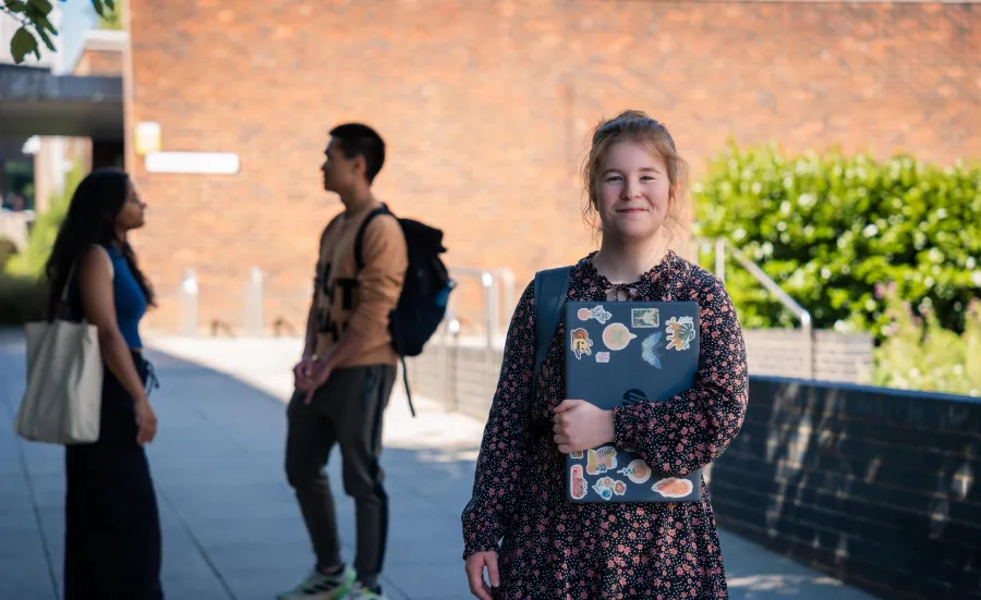 A student smiling while stood outside and holding a closed laptop. There are two more students talking to each other in the background.