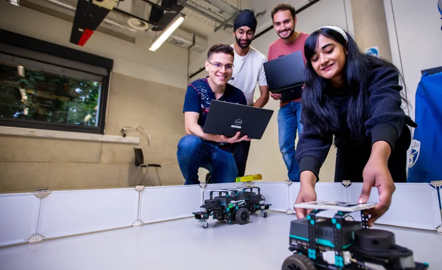 A group of 4 students are programming and watching 2 wheeled robot vehicles in a test area. Two of the students have laptops open while a third student is positioning one of the robotic vehicles.