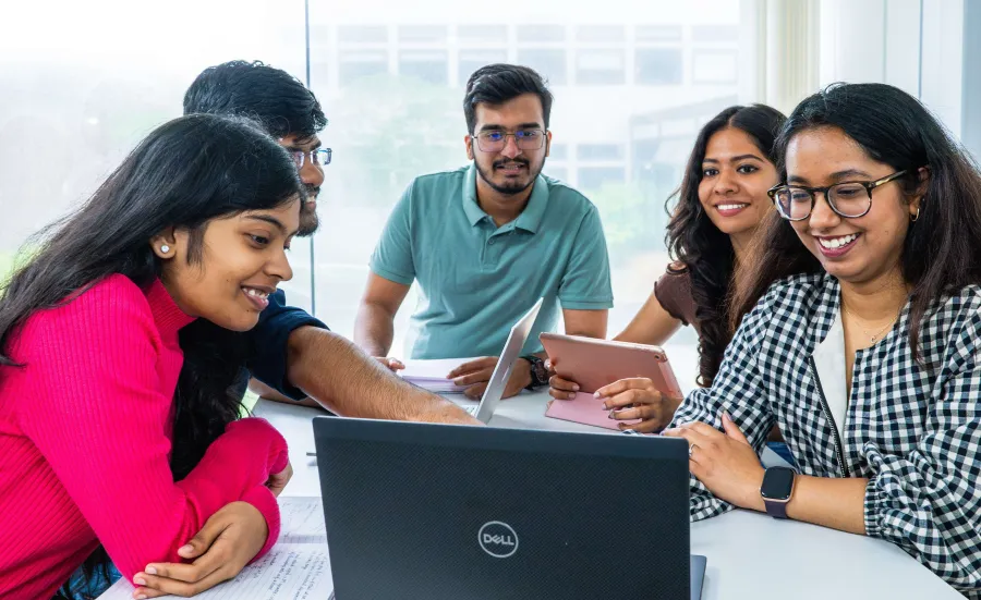 A group of university students work together at a table, smiling and using laptops and tablets.