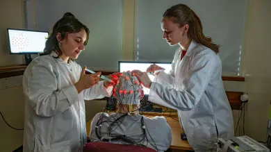 Two students adjust electrodes on a brain cap with red lights in a lab.