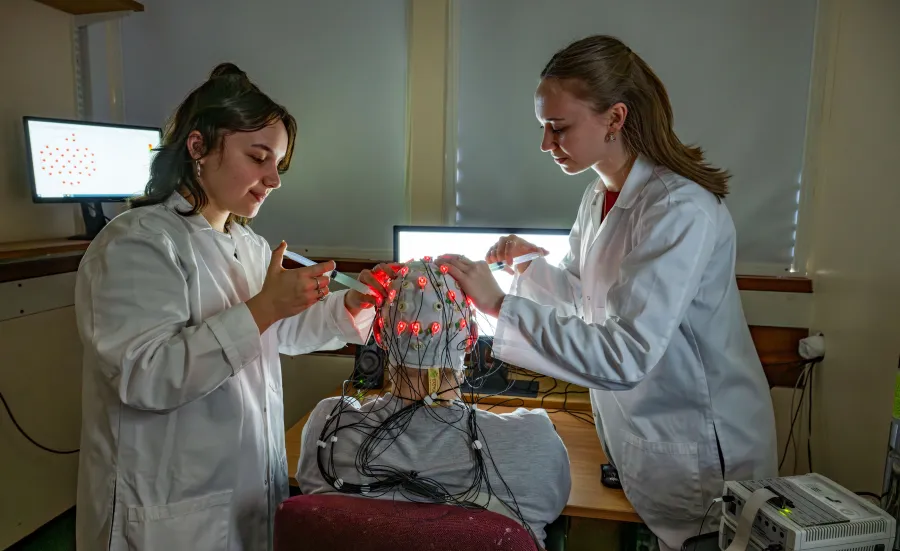 Two students adjust electrodes on a brain cap with red lights in a lab.